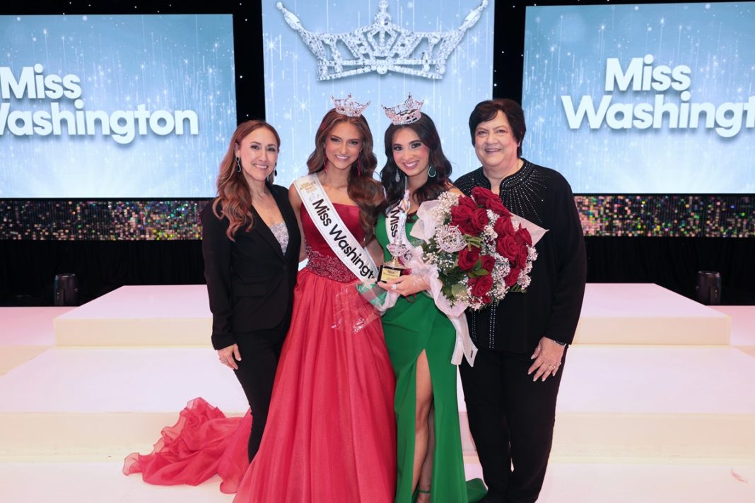Four women post on a stage. The two on either ends are in black pant suits, the one on the left middle in a red gown that says Miss Washington's Teen and the middle right in a green gown with a sash that says Miss Washington. A large screen with a crown on it and the Word's 'Miss Washington' on either side is in the background