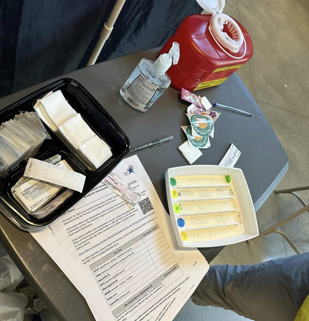 a table with paperwork, a sharps container, hand sanitizer, band aids, stickers, pens and other medical items
