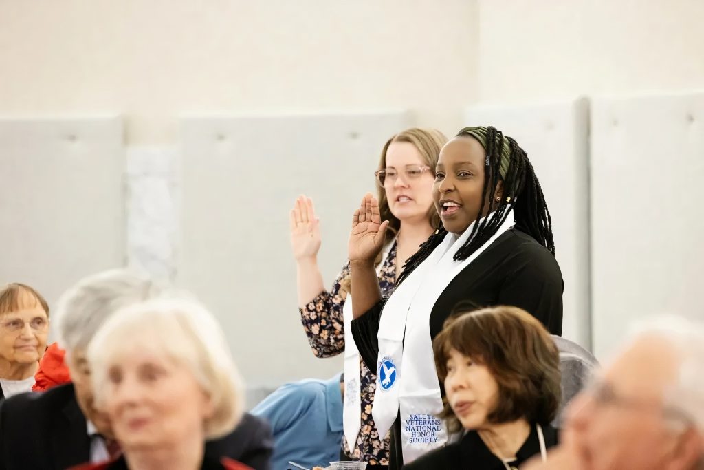 Two women with white stoles that say 'Salute Veteran National Honor Society' Stand up in a group of people with their right hands raised like they are taking a pledge