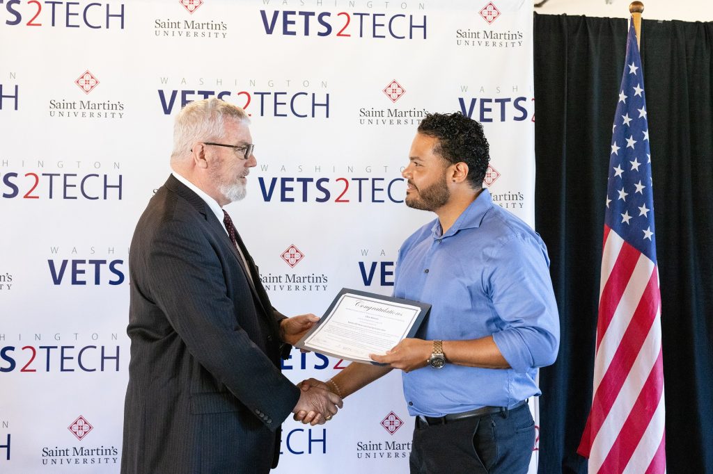 a man shakes hands with another man while handing him a certificate. They stand in front of a step-and-repeat that says 'Vets2Tech' and has the Saint Martin's University logo on it