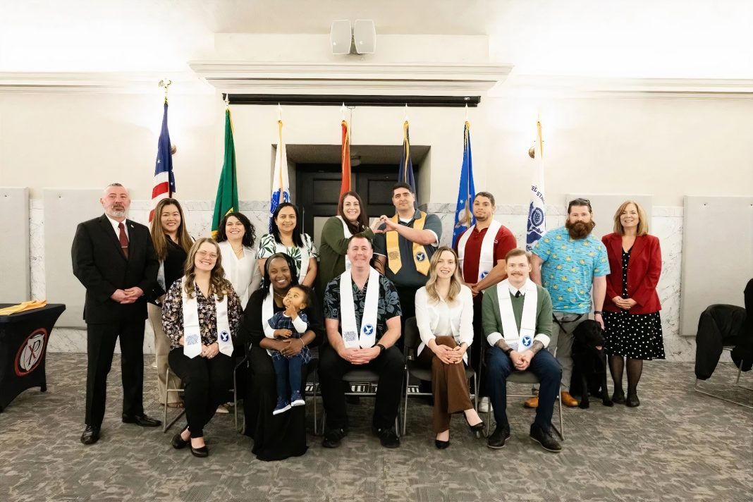 A group of veteran graduates, five sitting in chairs, one with a baby in her lap, 9 people standing behind them. 6 of the people have white graduation stoles. One has a yellow stole