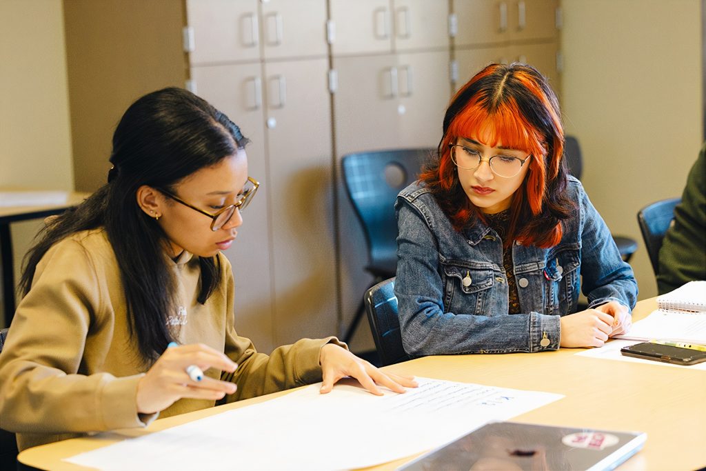Two women sit at a table together looking at some papers. One is holding a marker and has a hand on one piece of paper
