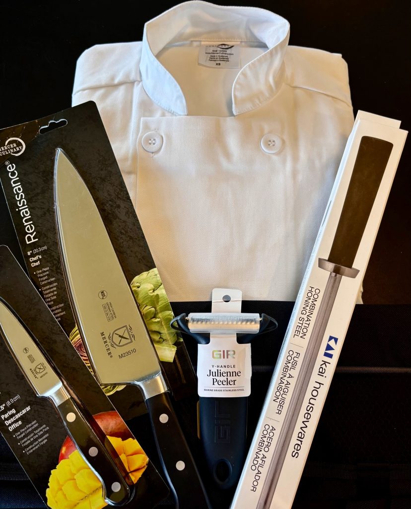 a chef's jacket, two knives, a julienne peeler and and another utensil laid out against a black background
