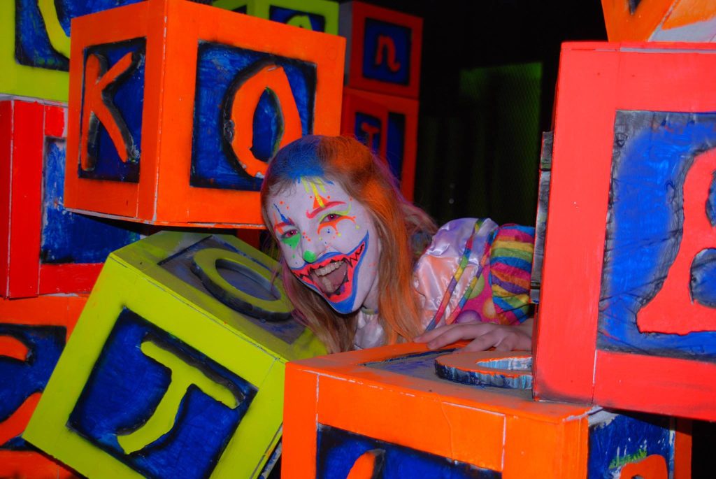 large florescent letter blocks are piled together with a person dressed as an evil clown peeking out from them.