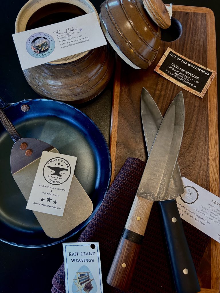 wood board with two knives on it, a crockery pot, sauce pan with a metal flipper and more on a table with business cards on top of them.