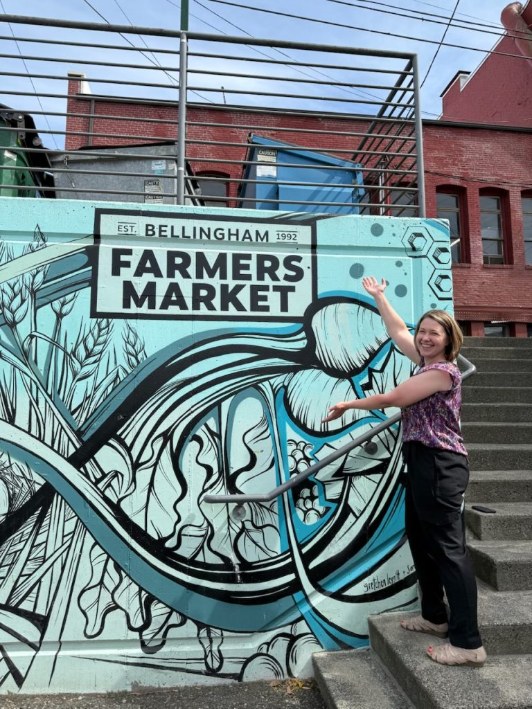 Juliana Venables posing by wall art on a concrete wall that says Bellingham Farmers Market