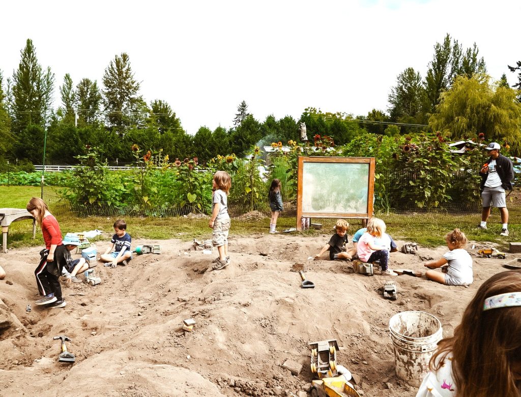 a bunch of young kid play in the dirt, a garden is behind them a large wooden sign