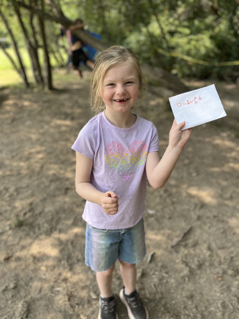 a girl standing outside with smiling and holding up a piece of paper with the name Olivia written on it and a crayon in her other hand