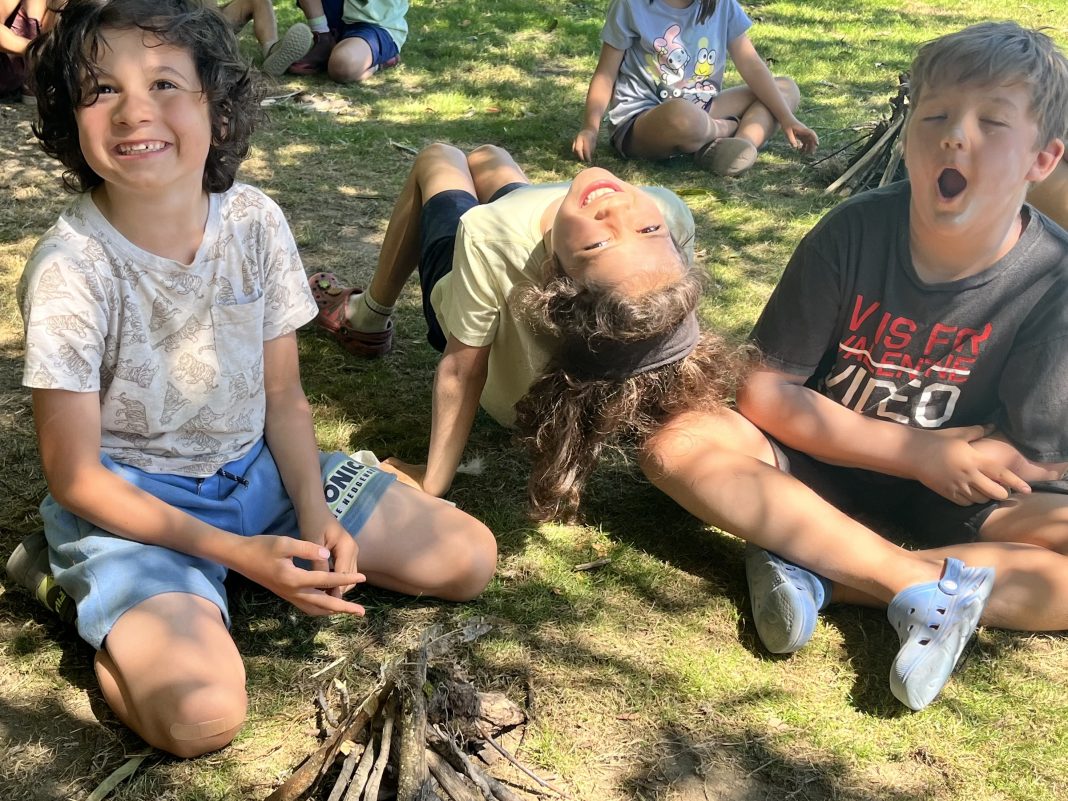 kids sit in the grass. One with a pile of sticks in front of her. Another is arching back with hands and feet beneath her, looking upside down at the camera