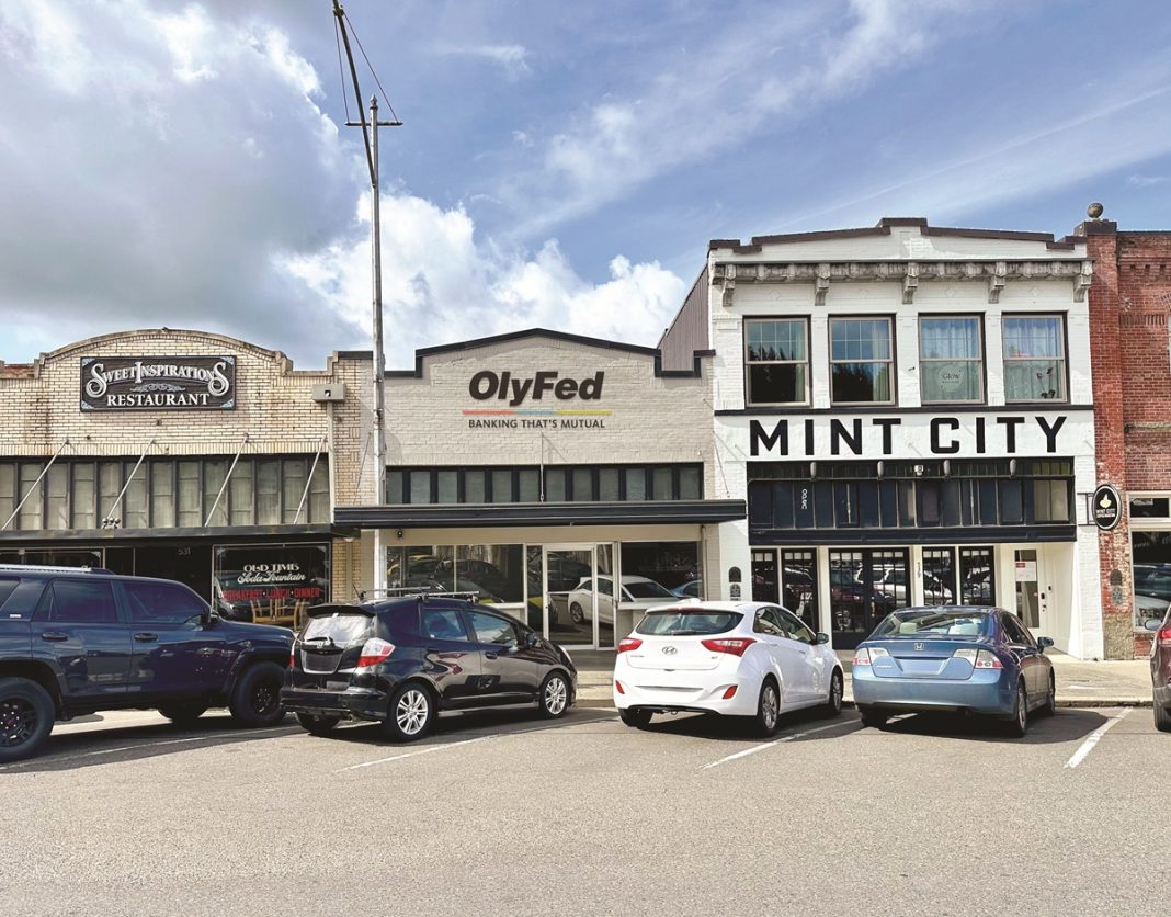 Row of store fronts with signs that say "Sweet Inspirations Restaurant," "OlyFed Banking that's mutual" and "Mint City" in downtown Chehalis. Cars are parked out front in angled parking spots.