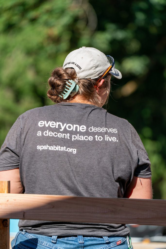 a woman with her back turned toward the camera with a grey shirt on that says "everyone deserves a decent place to live. spshabitat.org"